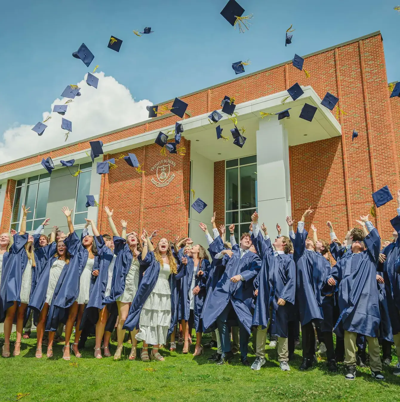 Exuberant high school graduates in blue gowns joyfully tossing their caps in the air in front of their school building.