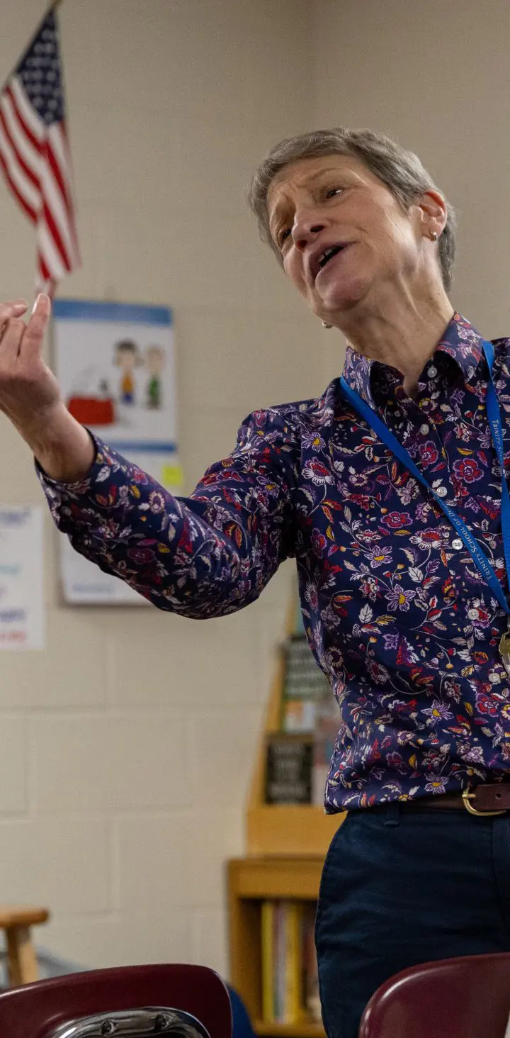 An animated female teacher explaining a concept with hand gestures in a classroom with students and educational posters in the background.