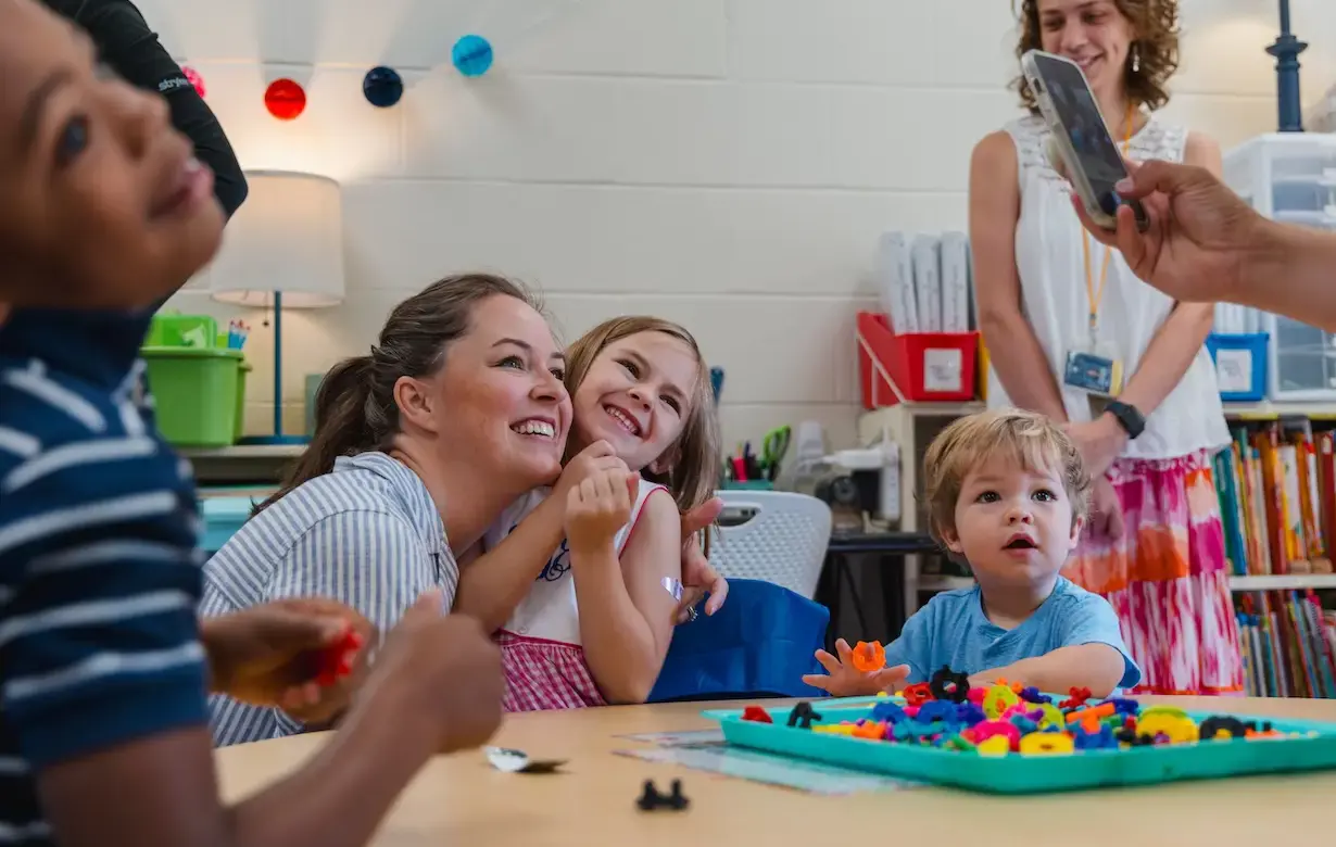 A mother embraces her daughter whilst sat at a table with other young children and a teacher in the background