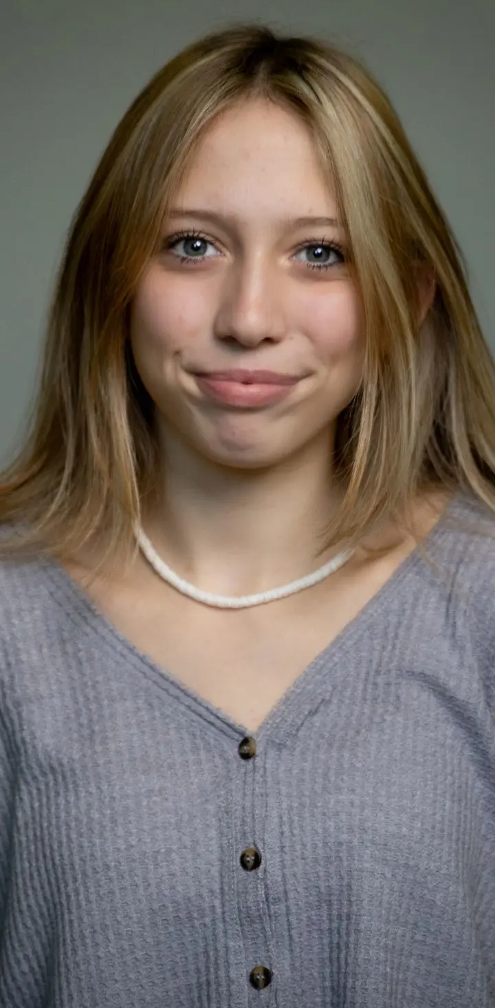 A young blonde girl in a grey cardigan smiling subtly at the camera.