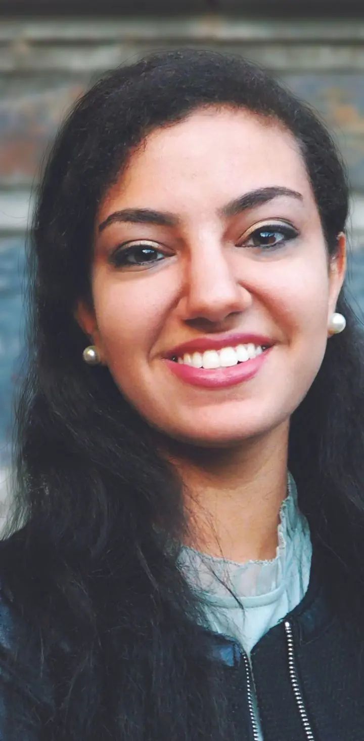 A smiling young woman with long dark hair wearing a leather jacket and a light-ruffled blouse.