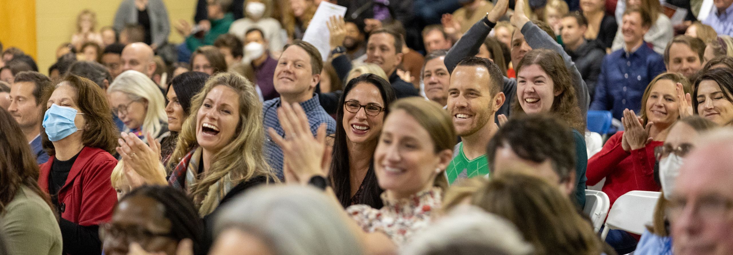 A diverse audience of adults smiling, clapping, and capturing moments with smartphones at an indoor school event. The atmosphere is lively and joyful, with a sense of community and engagement.