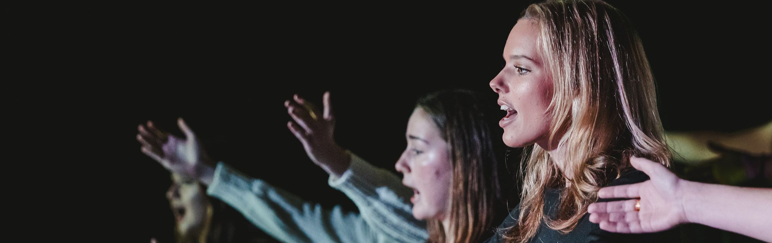 High school drama students emotively performing on stage, with arms extended and expressions full of intensity, during a dramatic evening theater production.