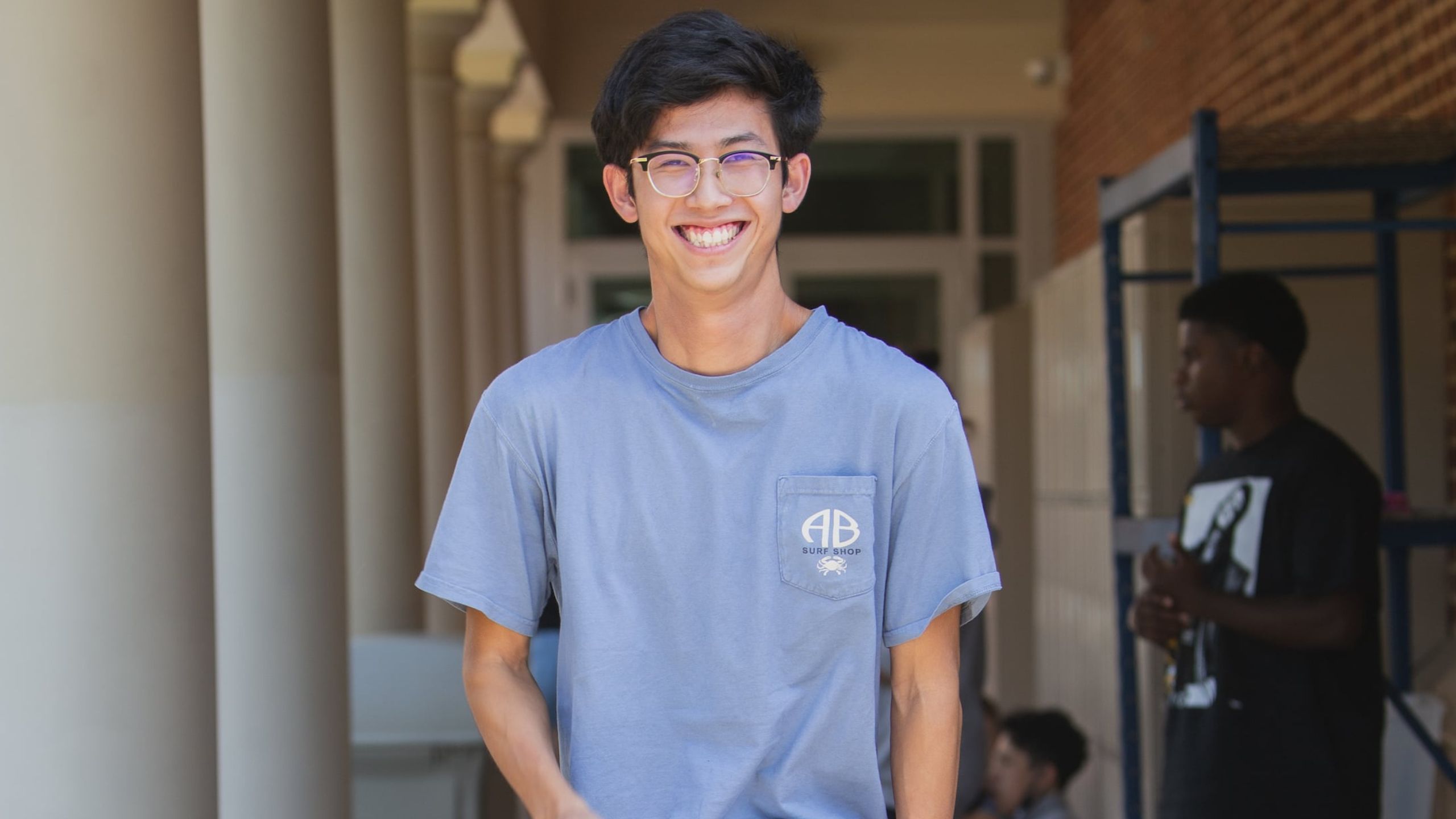 A cheerful teenage boy wearing glasses, a grey t-shirt, and khaki shorts stands in a school corridor with students sitting and standing in the background. He is smiling broadly towards the camera.