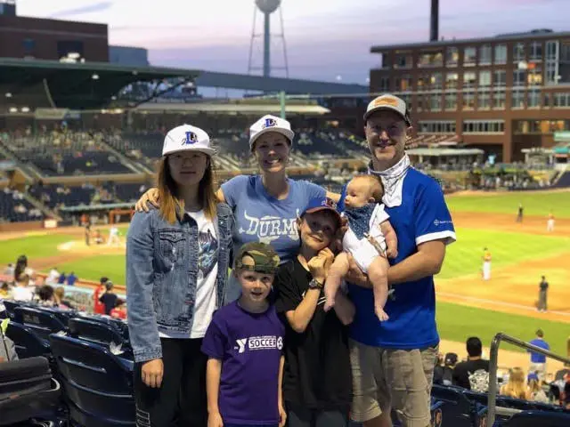 A family of five with two adults and three children posing for a photo at a baseball stadium during dusk. They are smiling and wearing casual sporting attire, with the baseball field and stadium seats in the background.