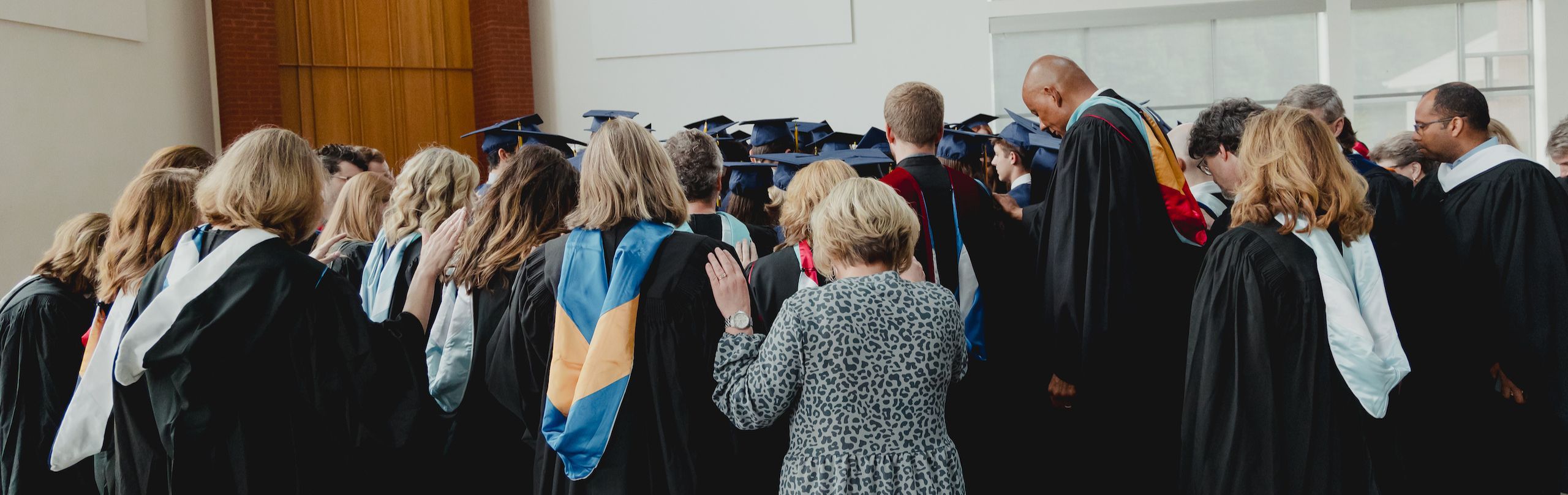 Group of graduating students in blue caps and gowns bowed in prayer, surrounded by faculty in academic regalia, in a ceremony hall with large windows and wooden accents.