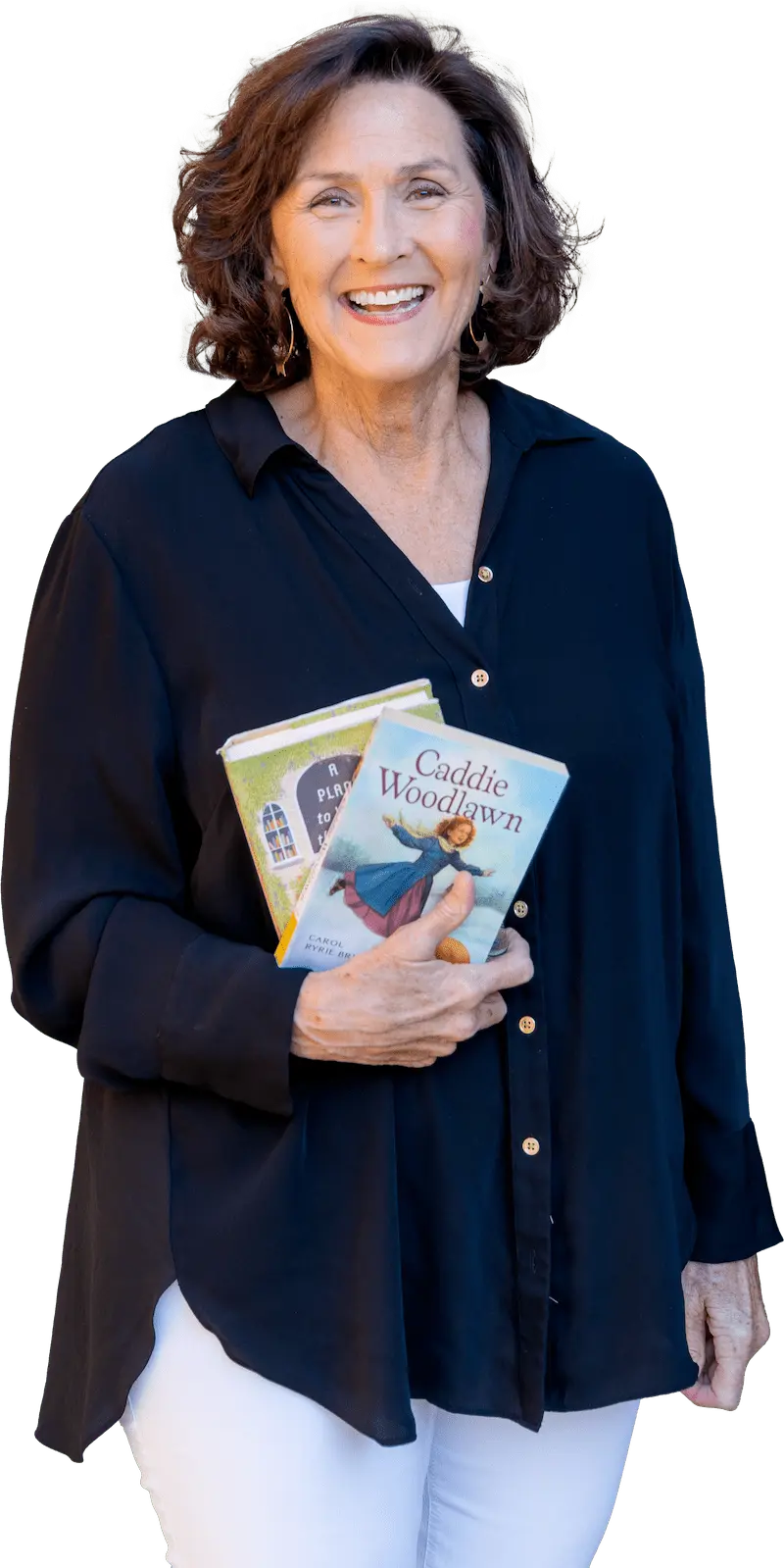 Cheerful woman with curly hair, wearing a navy blouse and white pants, holding two children's books, with a radiant smile