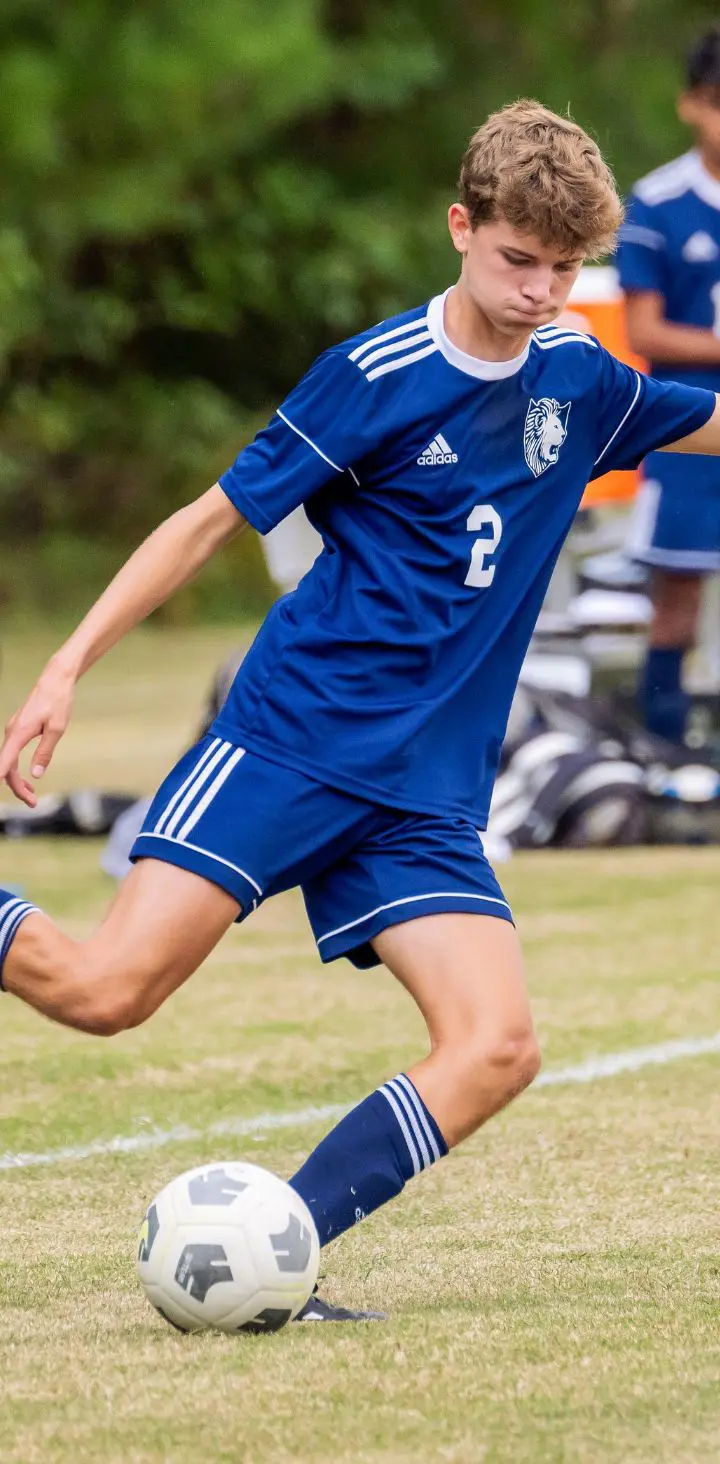 A focused young soccer player in a blue uniform performing a powerful kick during a match on a grassy field.