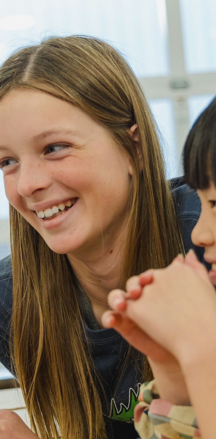 Three students are sharing a delightful moment over a learning activity; the focus is on a smiling girl with long hair, while her classmates look on with curiosity and joy in a bright classroom.